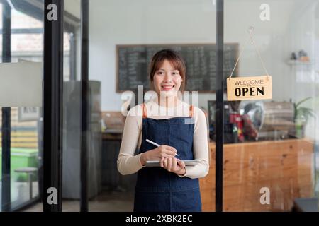 Junge Frau Cafe-Eigentümerin, die ein kleines Unternehmen gründet, lächelnder Unternehmer in Schürze hält Notebook, begrüßt Kunden mit offenem Schild, moderner Coffee Shop Stockfoto