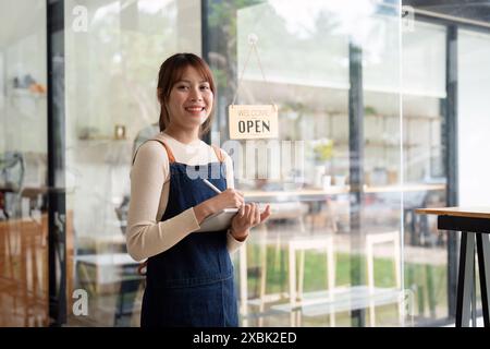 Junge Frau, die ihr eigenes kleines Unternehmen als Cafe-Besitzer gründet, lächelt und einen Notizblock in einem modernen Coffee Shop mit einem Willkommensschild auf dem hält Stockfoto