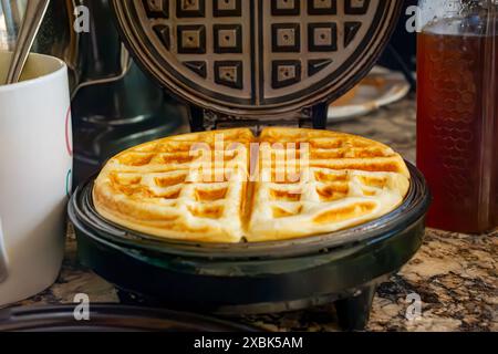 Auf dem Waffeleisen befindet sich eine fertige Waffel mit Teigbecher und Honig im Hintergrund. Stockfoto