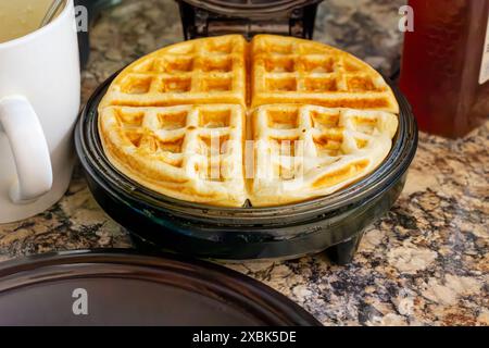 Auf dem Waffeleisen befindet sich eine fertige Waffel mit Teigbecher und Honig im Hintergrund. Stockfoto