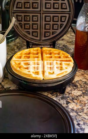 Auf dem Waffeleisen befindet sich eine fertige Waffel mit Teigbecher und Honig im Hintergrund. Stockfoto