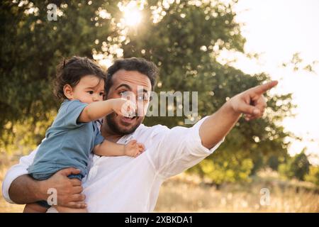 Ein einjähriger Junge, der von seinem Vater mit Bäumen und Bokeh im Hintergrund festgehalten wird, um einen herzerwärmenden Moment am Vatertag festzuhalten Stockfoto