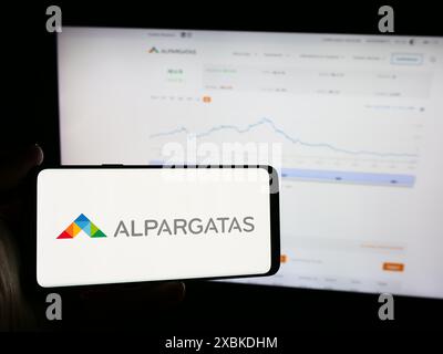 Person, die ein Mobiltelefon mit dem Logo des brasilianischen Schuhunternehmens Alpargatas SA vor der Business-Webseite hält. Schwerpunkt auf dem Display des Telefons. Stockfoto