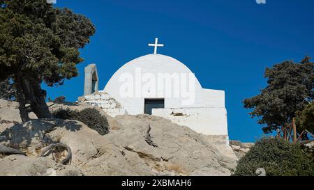 Weiße Kirche mit einem Kreuz auf einem felsigen Hügel, umgeben von Bäumen unter einem klaren blauen Himmel, Kapelle von Agios Panteleimon, Kastro Monolithou, Monolithos Stockfoto