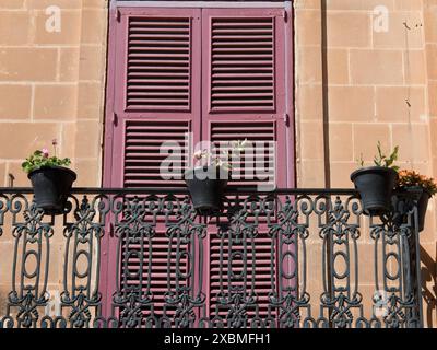 Ein Balkon mit schmiedeeisernen Geländern und rosa Fensterläden, dekoriert mit mehreren Blumentöpfen vor einer Sandsteinfassade, mdina, mittelmeer Stockfoto