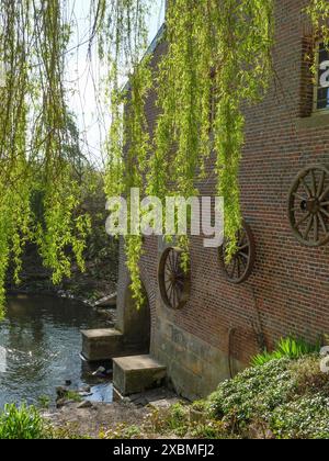 Ein Backsteinbau mit großen Holzrädern befindet sich am Ufer, umgeben von grünen Bäumen und überhängenden Ästen an einem sonnigen Frühlingstag, metelen Stockfoto