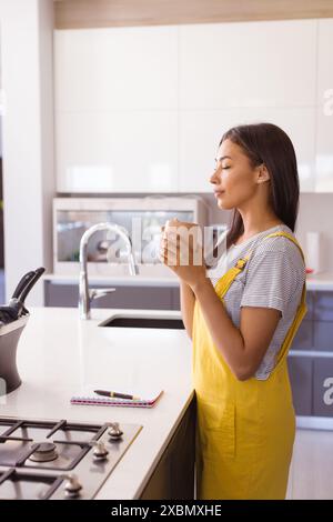 Glückliche Frau in gelber Latzhose, die einen Becher in einer modernen Küche hält Stockfoto