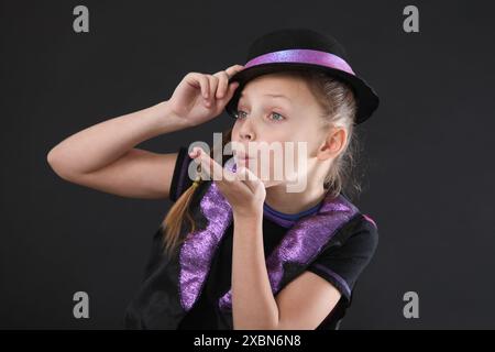 Porträt eines glücklichen Mädchens im Studio auf schwarzem Hintergrund, in einer schwarzen Weste und einem Hut gekleidet, das Gesichtsausdrücke macht und schauspielerische Fähigkeiten übt Stockfoto