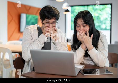 Zwei aufgeregte, schockierte junge asiatische Freunde bedecken ihren Mund und sind erstaunt über etwas auf einem Laptop-Bildschirm. Unerwartete gute Nachrichten, unglaublich Stockfoto