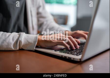 Ein Nahbild eines asiatischen Mannes in bequemer Kleidung, der an seinem Laptop arbeitet, auf der Tastatur des Laptops tippt, drinnen an einem Tisch arbeitet. Stockfoto