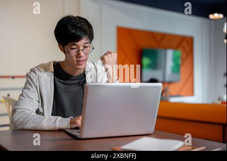 Ein junger asiatischer Mann in lässiger Kleidung und Brille zeigt seine geballte Faust, während er in einem Zimmer an seinem Laptop arbeitet. Menschen und Technologie Stockfoto