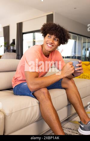 Glücklicher birassischer Mann, der auf der Couch sitzt und eine Kaffeetasse hält und lächelt Stockfoto