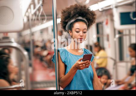 Junge schwarze Frau mit Afro-Frisur, die ihr Handy durchsucht, während sie sich im U-Bahn-Zug auf den Pol lehnt Stockfoto