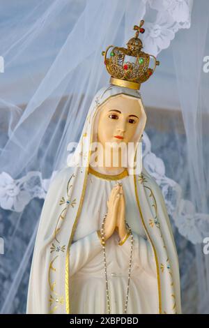 Statue der Jungfrau maria in friedlicher Pose, geschmückt mit einer goldenen Krone und einem weißen Schleier, mit einem Rosenkranz im Gebet Stockfoto