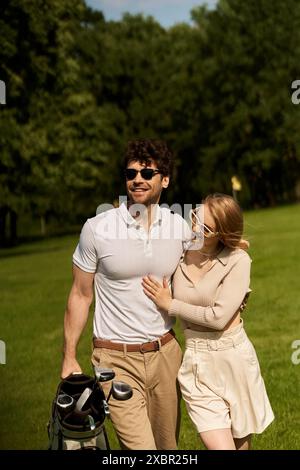 Ein stilvolles junges Paar in eleganter Kleidung, das gemütlich mit einer Golftasche in einer üppigen Parklandschaft spaziert. Stockfoto