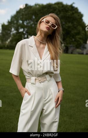 Eine stilvolle Frau im weißen Jumpsuit posiert auf einem weiten, grünen Feld, umgeben von Gelassenheit und Schönheit. Stockfoto