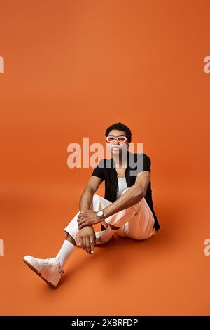 Hübscher Mann in Sonnenbrille, der vor leuchtendem orangefarbenem Hintergrund auf dem Boden sitzt. Stockfoto