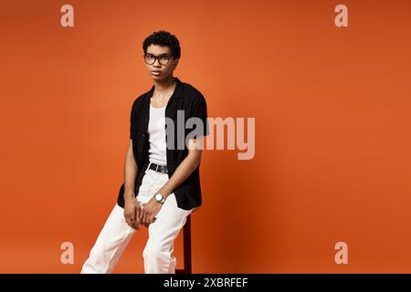 Ein hübscher afroamerikanischer Mann mit Sonnenbrille sitzt auf einem Hocker vor einer leuchtend orangefarbenen Wand. Stockfoto