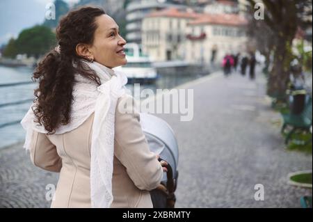 Eine fröhliche Frau spaziert mit ihrem Kinderwagen entlang eines ruhigen Flussweges. Die ruhige Umgebung trägt zur friedlichen und fröhlichen Atmosphäre bei Stockfoto