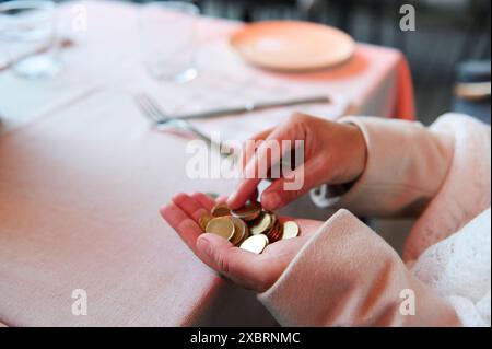 Eine Person zählt goldene Schokoladenmünzen in der Hand, während sie an einem Restauranttisch mit rosa Tischdecke, Gabel, Messer und Gläsern sitzt. Stockfoto