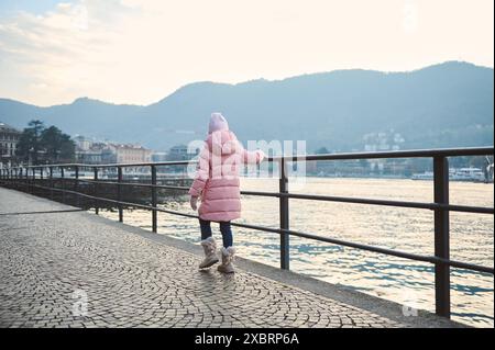 Ein junges Mädchen in einem rosa Wintermantel am See mit Bergen im Hintergrund. Friedliche Winterlandschaft mit ruhigem Wasser und fernen Bergen. Stockfoto