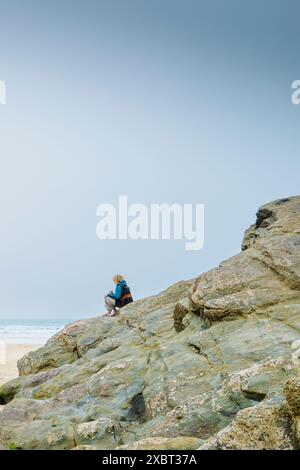 Eine Frau, die auf Felsen sitzt und das Meer am GT Western Great Western Beach an der Küste von Newquay in Cornwall in Großbritannien überblickt Stockfoto