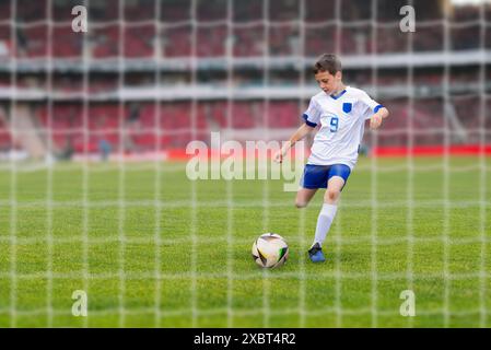 Junger Fußballspieler in einem weißen Kleid, der in einem modernen Stadion auf das Tor schießt. Blick hinter das Netz Stockfoto