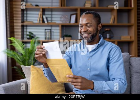 Glücklicher Mann, der einen Umschlag öffnet und einen Brief liest, während er auf einer Couch in einem komfortablen Wohnzimmer sitzt Stockfoto