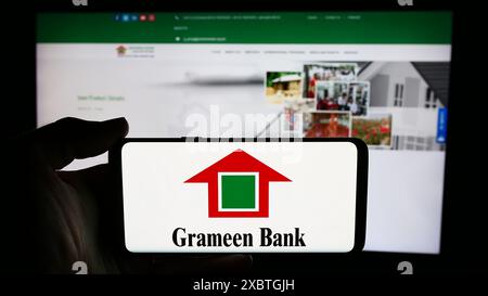 Person, die ein Mobiltelefon mit dem Logo des bangladeschischen Mikrofinanzunternehmens Grameen Bank vor der Business-Webseite hält. Schwerpunkt auf dem Display des Telefons. Stockfoto