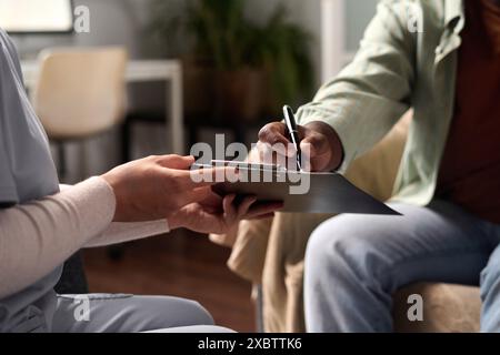 Seitliche Nahaufnahme eines nicht erkennbaren Schwarzen Mannes, der ein Dokument auf der Zwischenablage im Kopierraum der Klinik unterzeichnet Stockfoto