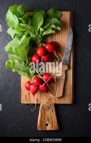Schneidebrett aus Holz mit zwei Haufen frischem rotem Bio-Rettich und Messer auf schwarzem Tisch. Draufsicht Stockfoto