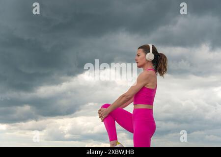 Sportliche Frau, die Beindehnungsübungen auf dem Wasser macht, trägt rosa Sportkleidung und Kopfhörer mit einem dramatischen Himmel. Stockfoto