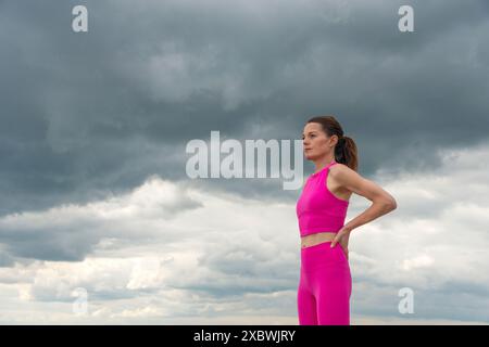 Sportliche Frau, die mit Händen auf den Hüften ruht und pinkfarbene Sportbekleidung vor einem dramatischen Himmel trägt. Stockfoto