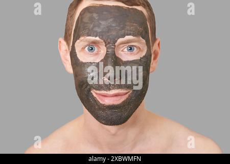 Schwarze Tonmaske auf dem Männergesicht. Kosmetologie Mann Porträt mit Schlamm gesundes Peeling. Kohle Natur Öko Masque. Dermatologisches Holzkohleprodukt. Häusliche Hautpflege. Hautpflege-Männerbehandlung. Grau Stockfoto