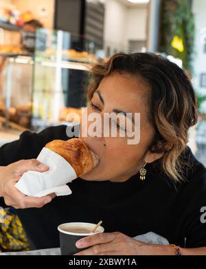 Eine hübsche hispanische Frau genießt ihr Croissant, indem sie es in ihren Kaffee taucht, während sie in einer Bäckerei in Paris sitzt. Stockfoto