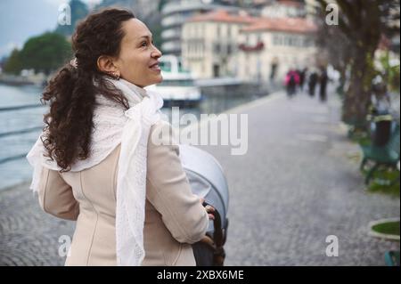 Eine fröhliche Frau spaziert mit ihrem Kinderwagen entlang eines ruhigen Flussweges. Die ruhige Umgebung trägt zur friedlichen und fröhlichen Atmosphäre bei Stockfoto