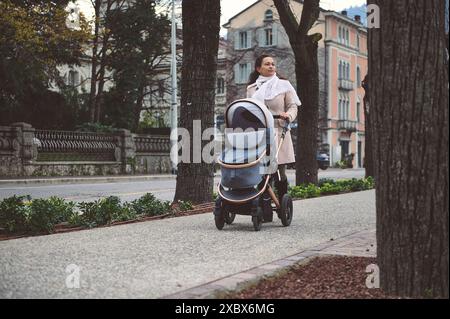 Eine junge Mutter macht in der Herbstsaison einen gemütlichen Spaziergang mit einem Kinderwagen auf einem Bürgersteig in der Stadt und genießt die ruhige städtische Umgebung. Stockfoto