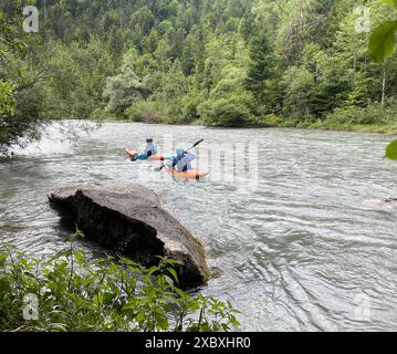 Zwei Kajakfahrer auf einem Fluss, Blaue Helme und Sicherheitsausrüstung, Orangene und blaue Kajaks, Dichte Vegetation und Bäume, großer Felsvorsprung im Vordergrund, Ruhiges Wasser mit leichten Strömungen, Dichter Wald im Hintergrund, Fluss umgeben von üppigem Grün, klar erkennbarer Felsvorsprung, Natürliche unberührte Landschaft, Outdoor-Aktivität, Abenteuer, Naturerlebnis, Wassersport, Erholung, Entspannung, Freizeit, Kajakfahren, Wildwasser, Paddeln, Sport in der Natur. *** Zwei Kajakfahrer auf einem Fluss, Blauhelme und Sicherheitsausrüstung, orange und blaue Kajaks, dichte Vegetation und Bäume, großer roc Stockfoto