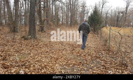 Ein einsamer älterer Mann geht einen mit Blättern bedeckten Weg durch den Wald. Ein alter, baufälliger Hirschstand ist links zu sehen. Stockfoto