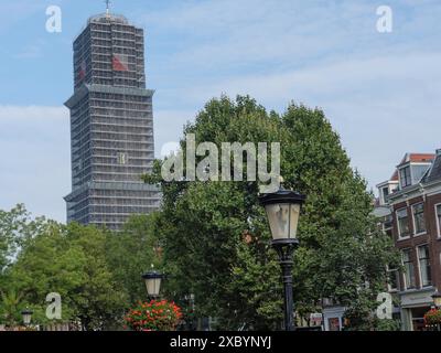 Blick auf die Straße mit gepflanzten Laternen und einem hohen Turm mit Gerüsten, gesäumt von Bäumen und Häusern unter bewölktem Himmel, utrecht, Niederlande Stockfoto