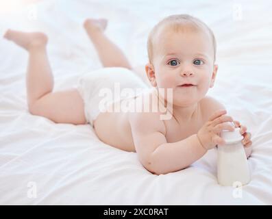 Baby, Porträt und Spiel auf dem Bett mit Flasche für Milchernährung, kindliche Entwicklung und gesunde Knochen zu Hause. Junge Säuglinge, Neugeborene und flüssiges Kalzium Stockfoto