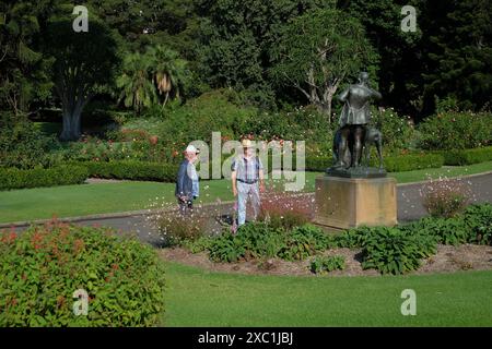 Zwei Männer sehen die Huntsman and Dogs Statue in den Royal Botanical Gardens Sydney mit grünen Rasenflächen und etablierten Bäumen und Grenzplantagen Stockfoto
