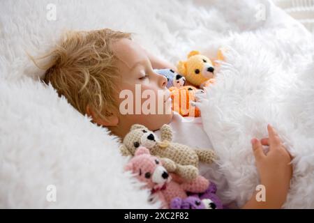 Kleines Kleinkind, niedlicher blonder Junge, der mit vielen Teddybären schläft, handgemachtes Amigurumi-Spielzeug zu Hause im Bett Stockfoto