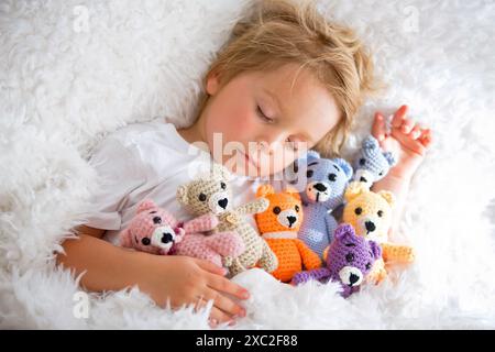 Kleines Kleinkind, niedlicher blonder Junge, der mit vielen Teddybären schläft, handgemachtes Amigurumi-Spielzeug zu Hause im Bett Stockfoto