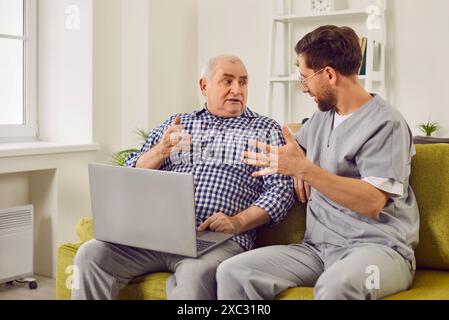 Senior Mann und junge Pflegekraft sitzen auf der Couch, reden miteinander und benutzen Laptop Stockfoto