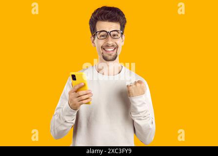 Ein staunender Mann in beigem Sweatshirt zeigt ja-Geste, der das Smartphone auf gelbem Hintergrund hält. Stockfoto