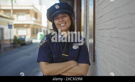 Eine lächelnde hispanische Polizistin mit überkreuzten Armen posiert selbstbewusst auf einer städtischen Straße. Stockfoto