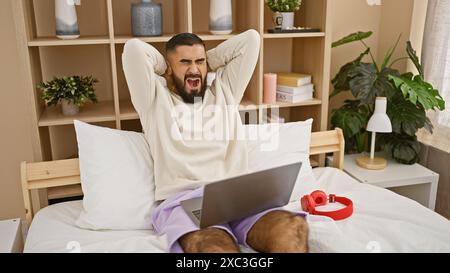 Ein gähnender Mann mit einem Bart in lässiger Kleidung, der sich auf einem Bett mit Laptop in einem gemütlichen Schlafzimmer ausbreitet. Stockfoto