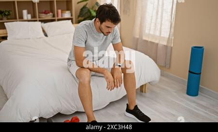 Nachdenklicher junger Mann, der auf einem Bett in einem Schlafzimmer mit Trainingsausrüstung sitzt und über seine Fitness-Routine nachdenkt. Stockfoto