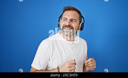Lächelnder reifer hispanischer Mann, der draußen Kopfhörer vor blauem Hintergrund trägt. Stockfoto